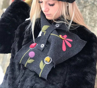 A woman wearing a black felt hat and matching black felt scarf with colorful embroidered flowers and a decorative band of red, yellow, and green patches, photographed outdoors against a stone wall.